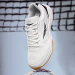 White Redtape men's badminton shoes with memory foam, black accents, and gum sole, hovering over a racket on a blurred court.